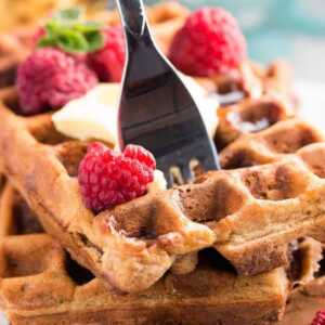 fork stuck in a stack of waffles topped with butter and raspberries