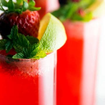 close up photo of champagne glasses filled with strawberry cocktail and garnished with lime, mint and fresh strawberry