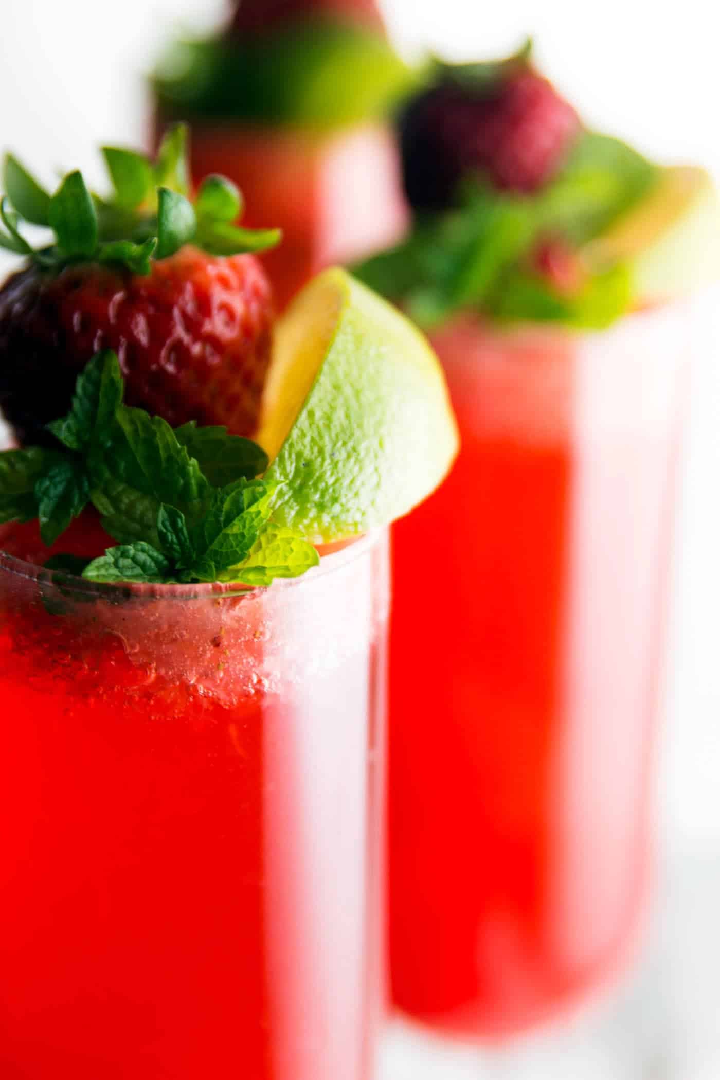 close up photo of champagne glasses filled with strawberry cocktail and garnished with lime, mint and fresh strawberry