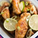 white enamel dish filled with crispy chicken wings, lime slices and chopped green onion on pink napkin