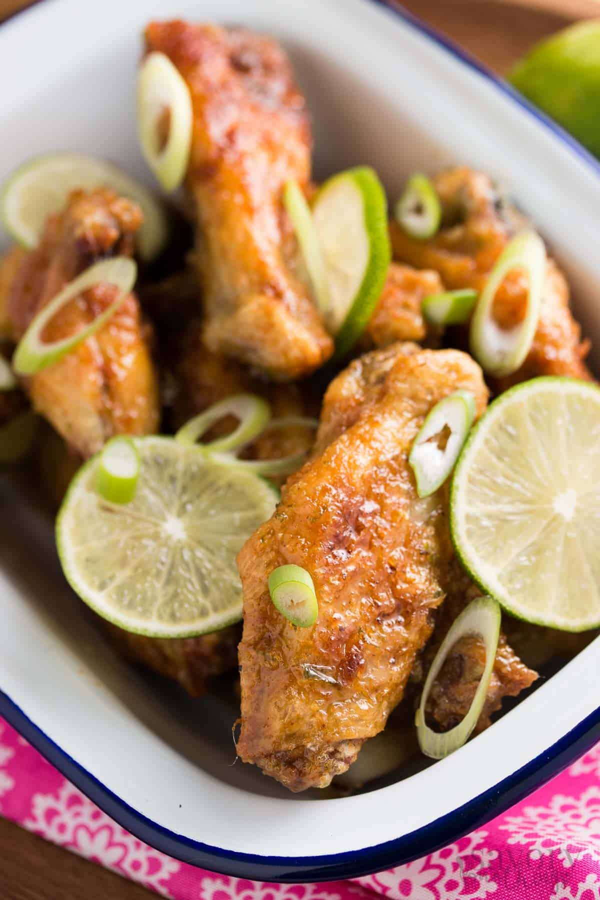 white enamel dish filled with crispy chicken wings, lime slices and chopped green onion on pink napkin