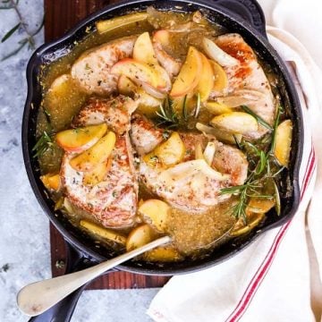 cast iron skillet filled with pork chops and apples, on a wooden board next to linen