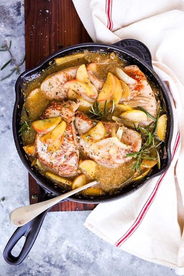 cast iron skillet filled with pork chops and apples, on a wooden board next to linen