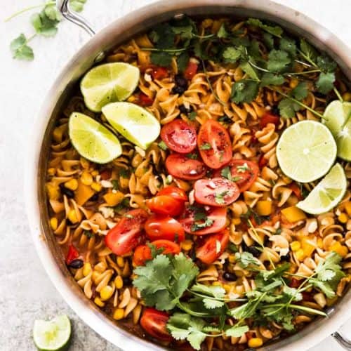 overhead photo of skillet with enchilada pasta, topped with cilantro, limes and tomatoes