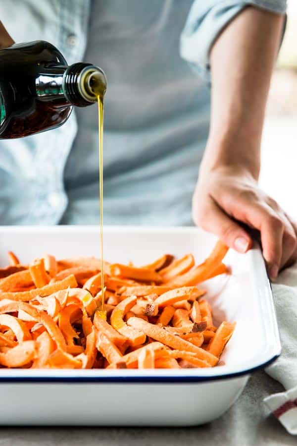 woman in chambray shirt drizzling olive oil over unbaked sweet potato fries in white enamel dish