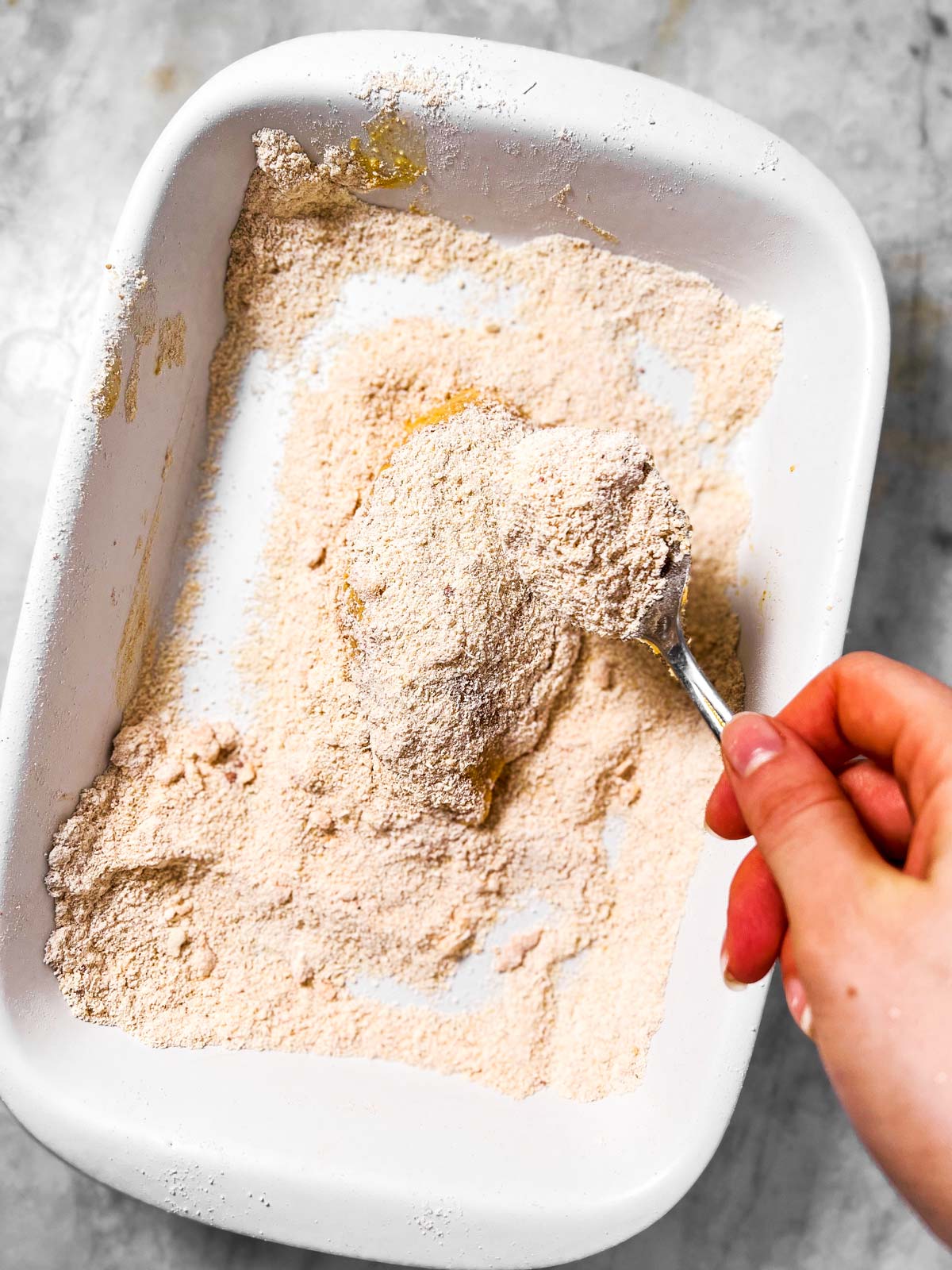 female hand spooning breading mix on chicken breast in white dish