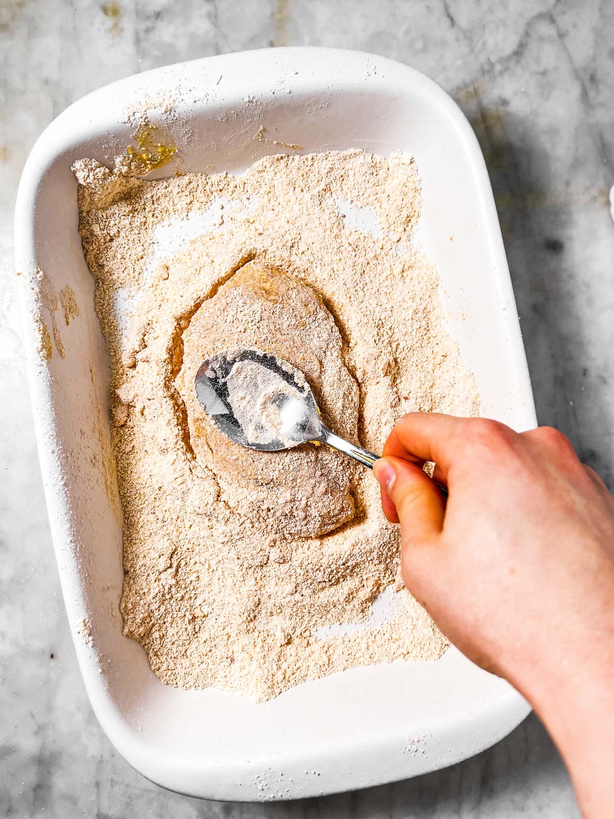 female hand using tablespoon to pound breaded chicken breast in white dish