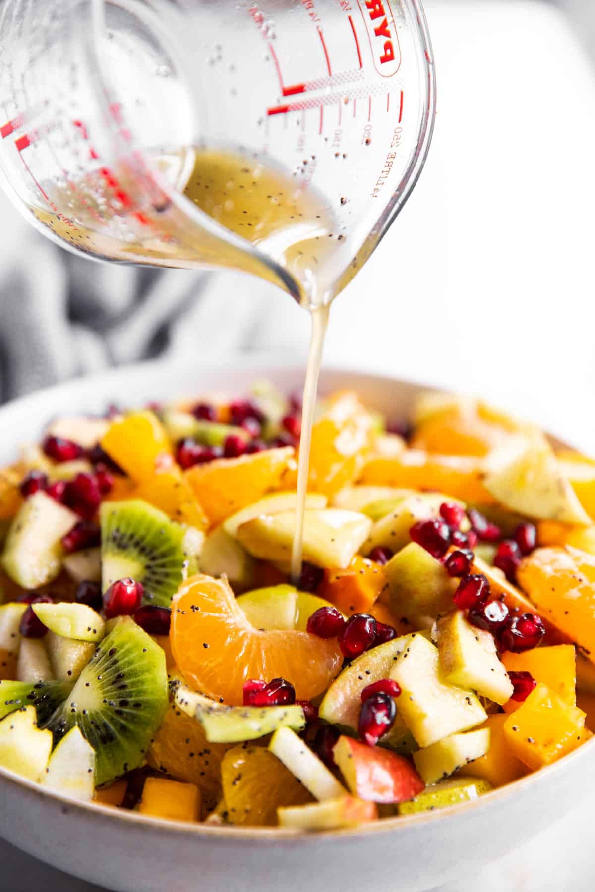poppy seed dressing pouring from a glass measuring jug over fruit salad