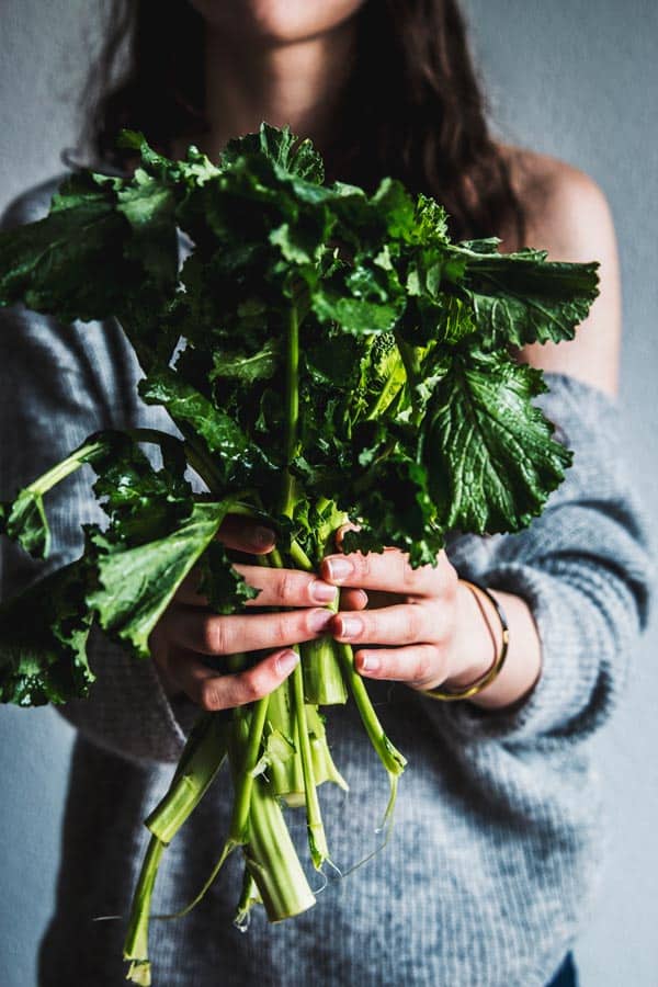 Woman in grey knit pullover, holding stems of broccoli rabe.