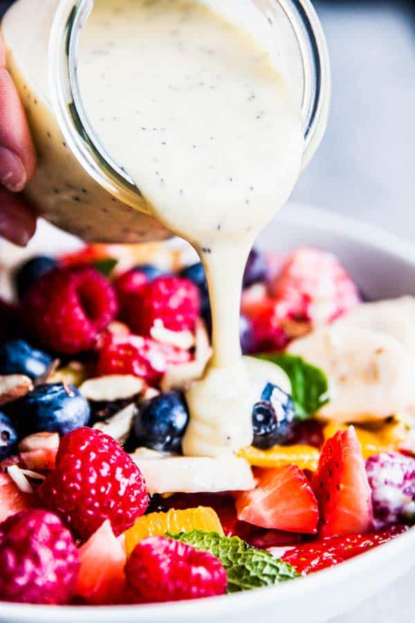 Pouring creamy poppy seed fruit salad dressing over fruit salad.
