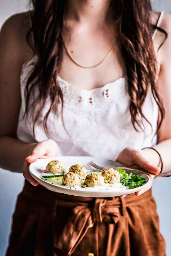 Woman in a white top and tan skirt holding a plate of greek turkey meatballs. 