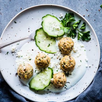 Greek turkey meatballs on a white plate with yogurt, cucumber slices and a fork. Black napkin next to it.