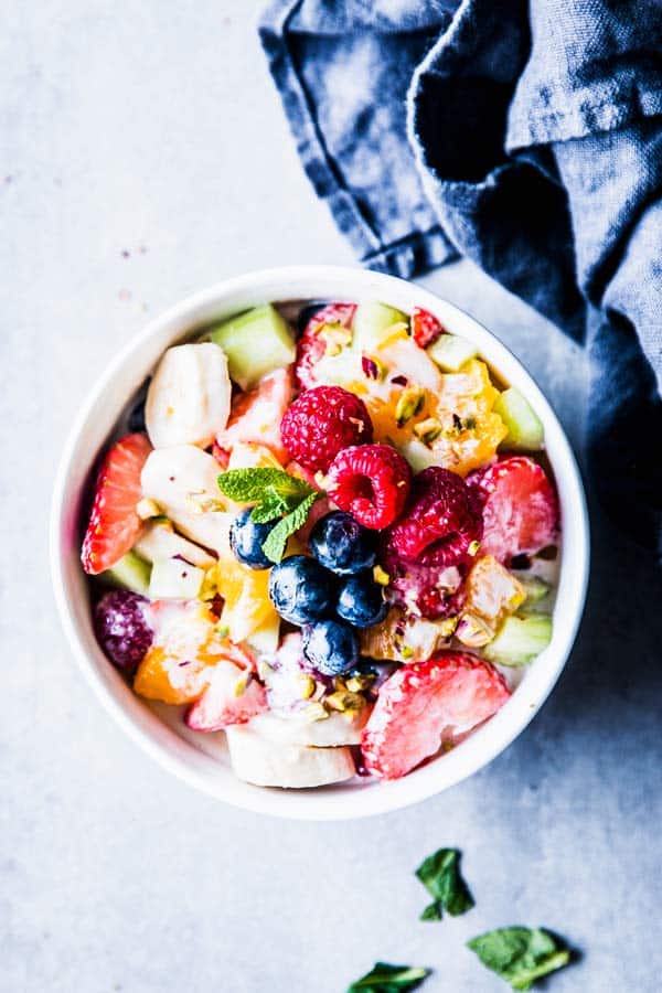 Greek Yogurt Fruit Salad in a white bowl with mint, next to a dark napkin.