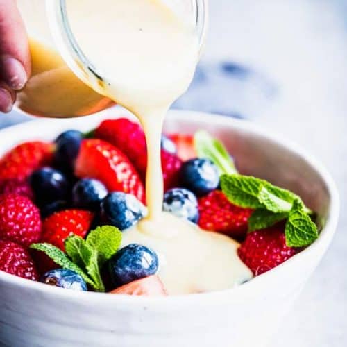 Pouring Creamy Fruit Salad Dressing over berry fruit salad in a white bowl.