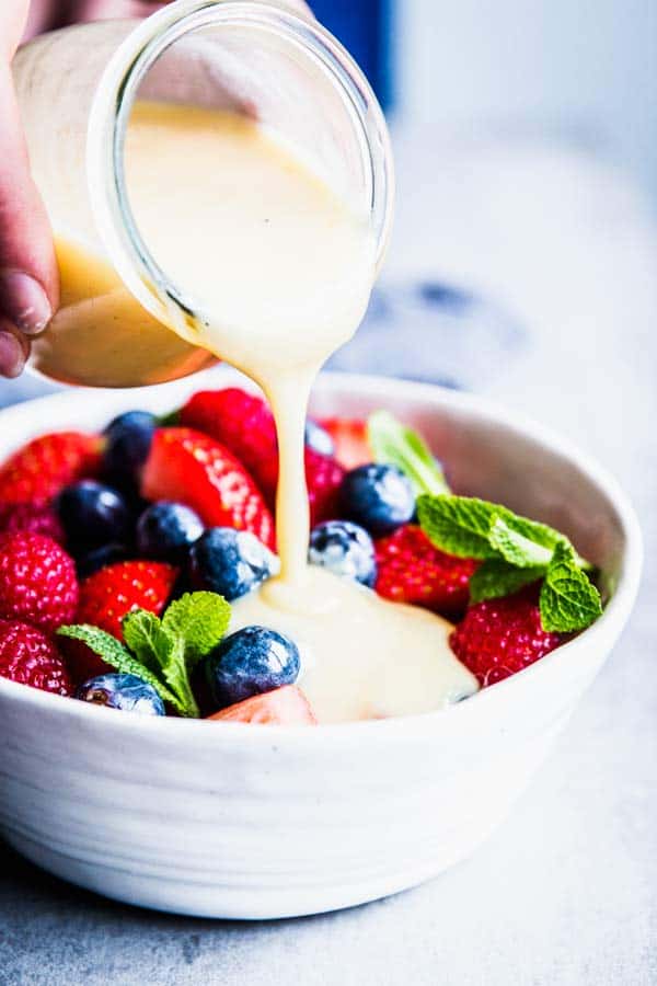Pouring Creamy Fruit Salad Dressing over berry fruit salad in a white bowl.