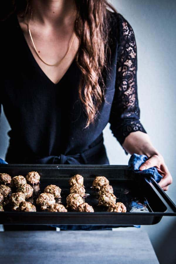 Woman in a black jumpsuit, holding a sheet pan with baked meatballs.