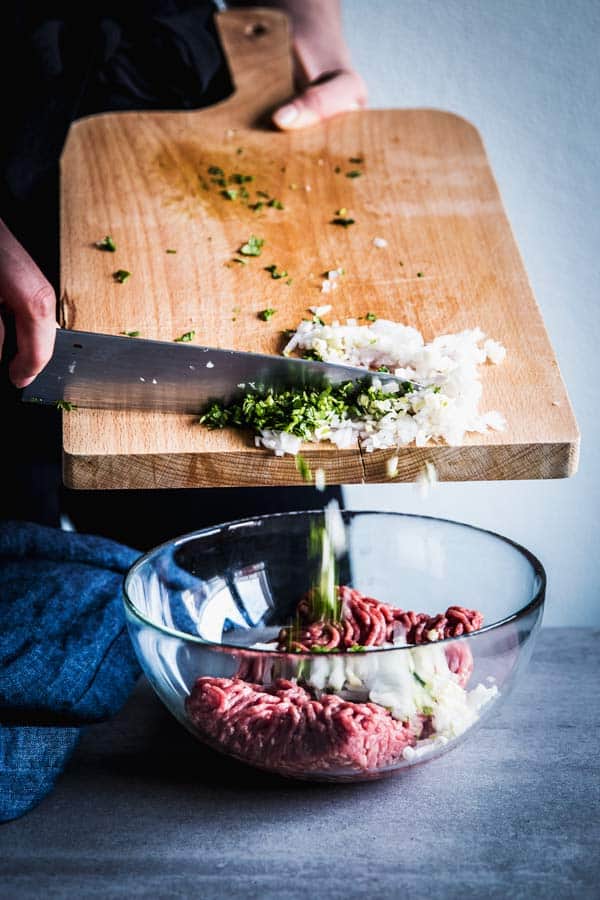 Woman pushing chopped cilantro, onion and garlic off a wooden chopping board into a glass bowl with ground beef.