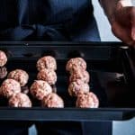 Woman in a black jumpsuit, holding a sheet pan full of homemade meatballs. Close up.