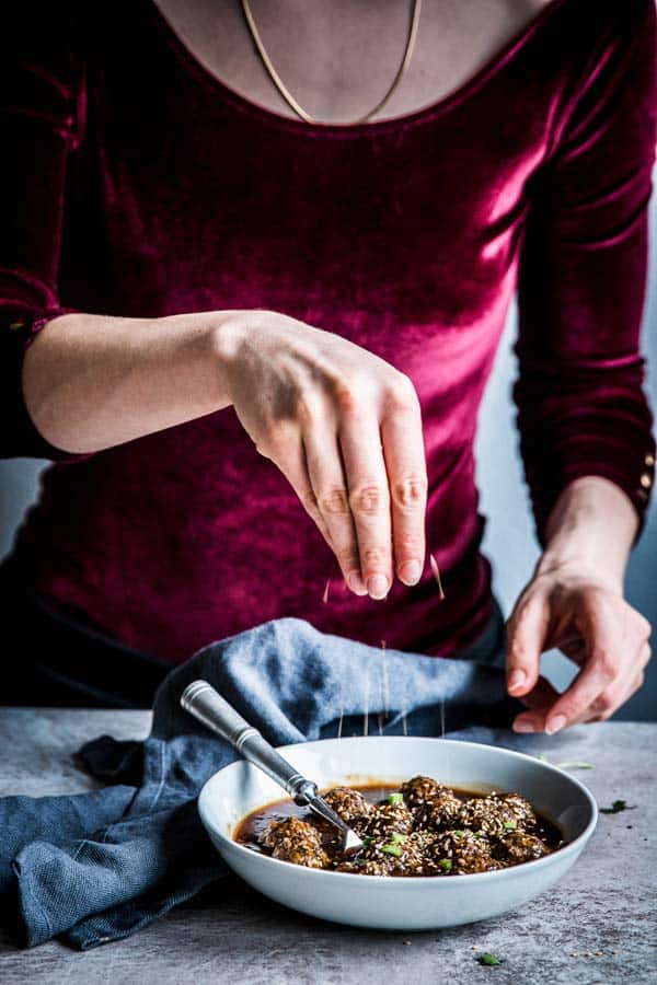 Woman in a red velvet top sprinkling sesame seeds over Korean BBQ Meatballs.