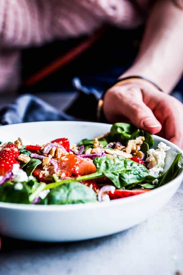 Woman placing a white bowl with Spinach Strawberry Walnut Salad on the table.
