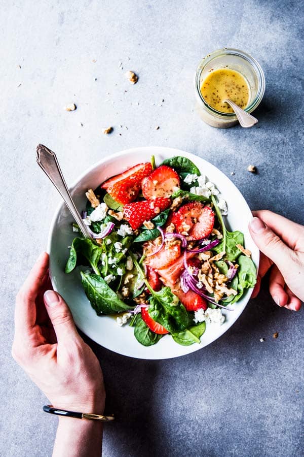 Hands placing a white bowl with spinach strawberry walnut salad on the table.