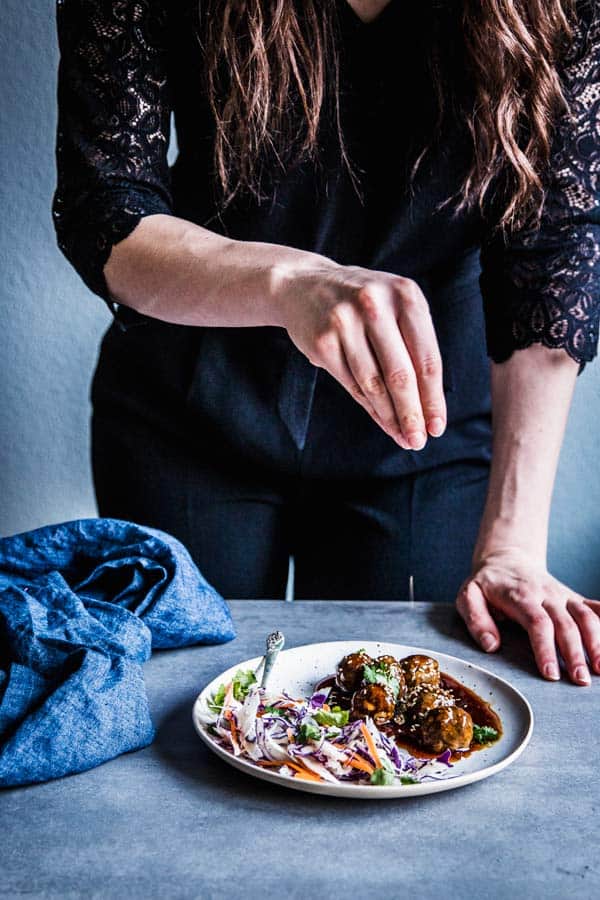 Woman sprinkling sesame seeds on a plate of teriyaki meatballs and coleslaw.