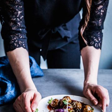 Woman in a black jumpsuit, serving a plate of teriyaki meatballs and coleslaw.