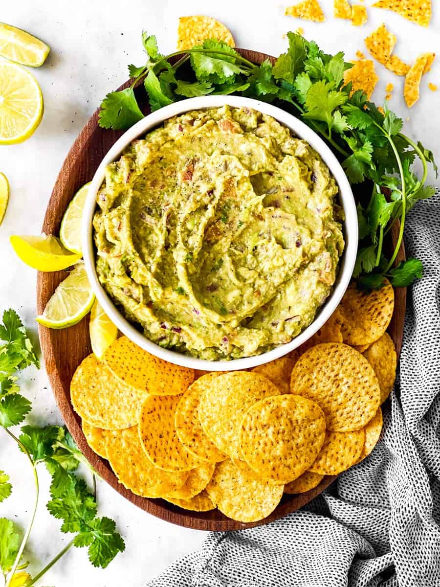 overhead view of wooden platter with bowl of guacamole, chips and cilantro