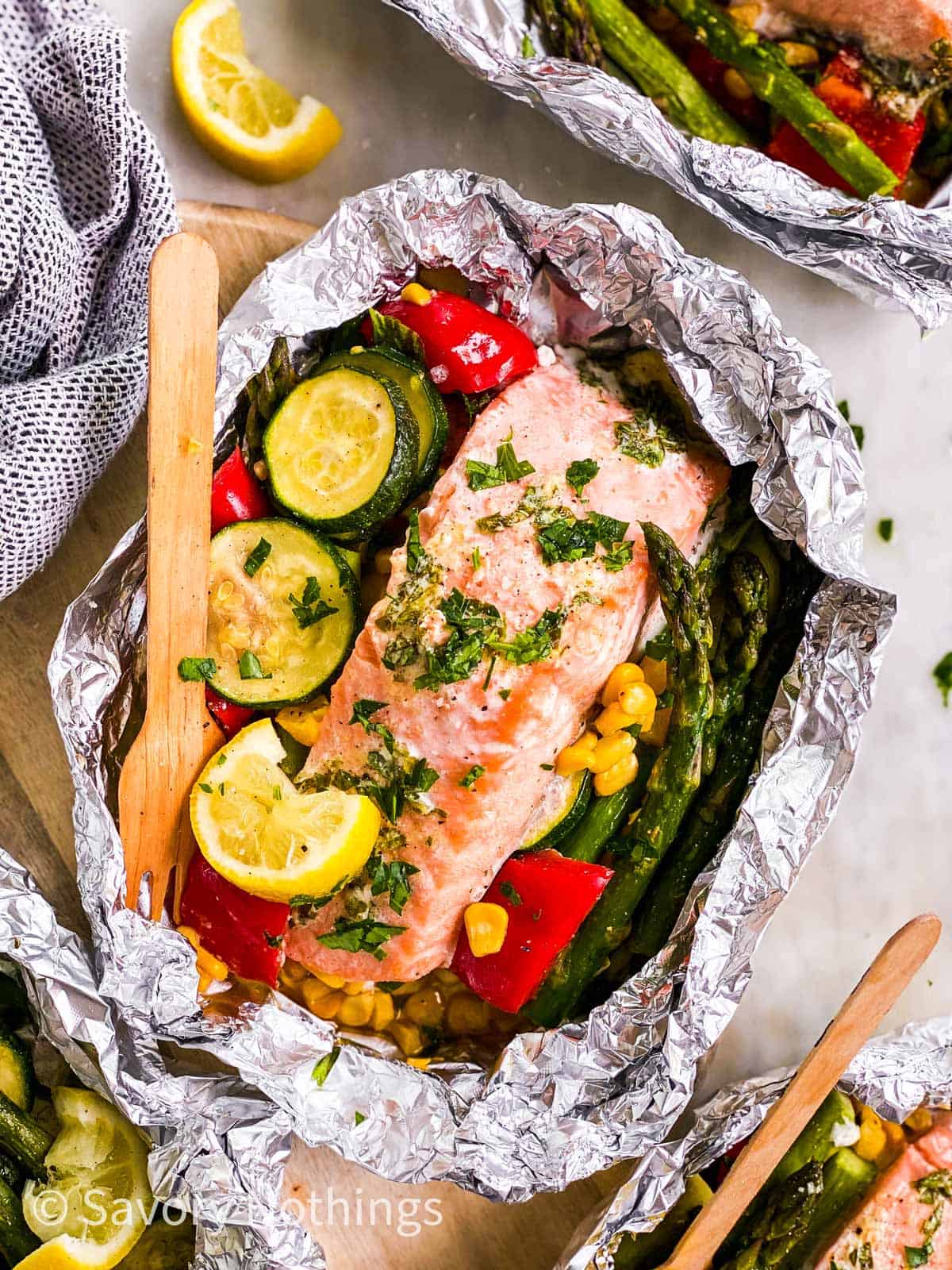 overhead view of salmon fillet in opened foil packet over bed of summer vegetables, with wooden fork stuck in vegetables