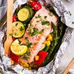 overhead view of salmon fillet in opened foil packet over bed of summer vegetables, with wooden fork stuck in vegetables