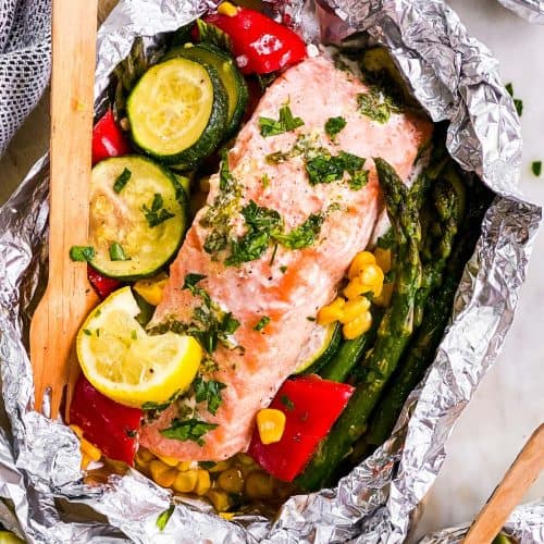 overhead view of salmon fillet in opened foil packet over bed of summer vegetables, with wooden fork stuck in vegetables
