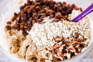 raisins, pecans and rolled oats on top of cookie dough in a glass bowl, with a purple spatula sticking out