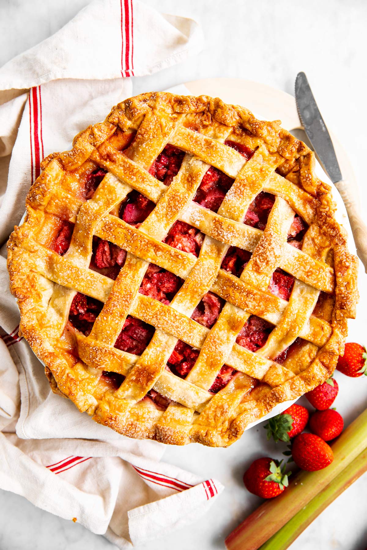 overhead view of strawberry rhubarb pie with dish towel and fresh fruit next to it