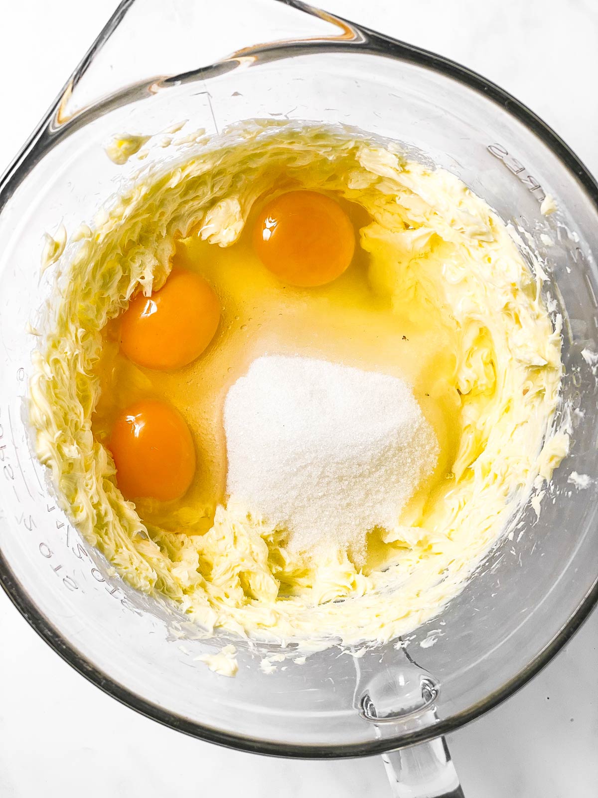 three cracked eggs and a pile of sugar o top of creamed butter in glass bowl