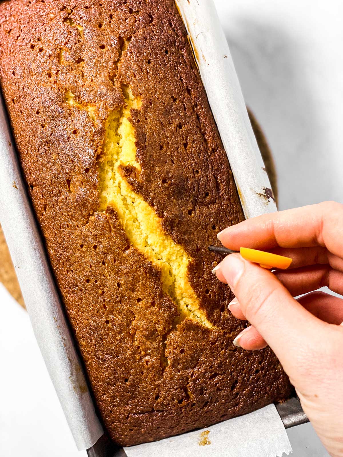 female hand using metal skewer to poke hole into lemon loaf cake