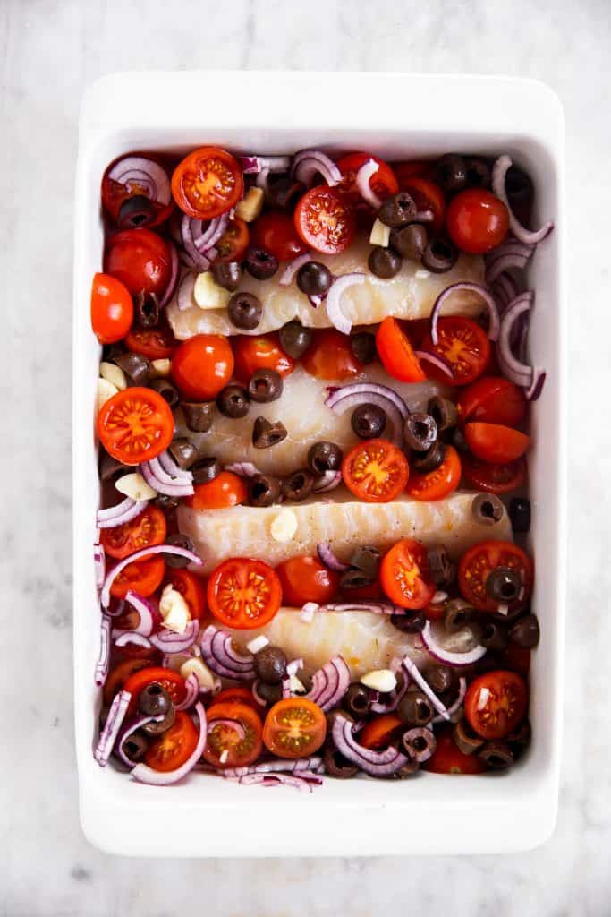 four cod fillets in white casserole dish with tomatoes, red onion, garlic and olives