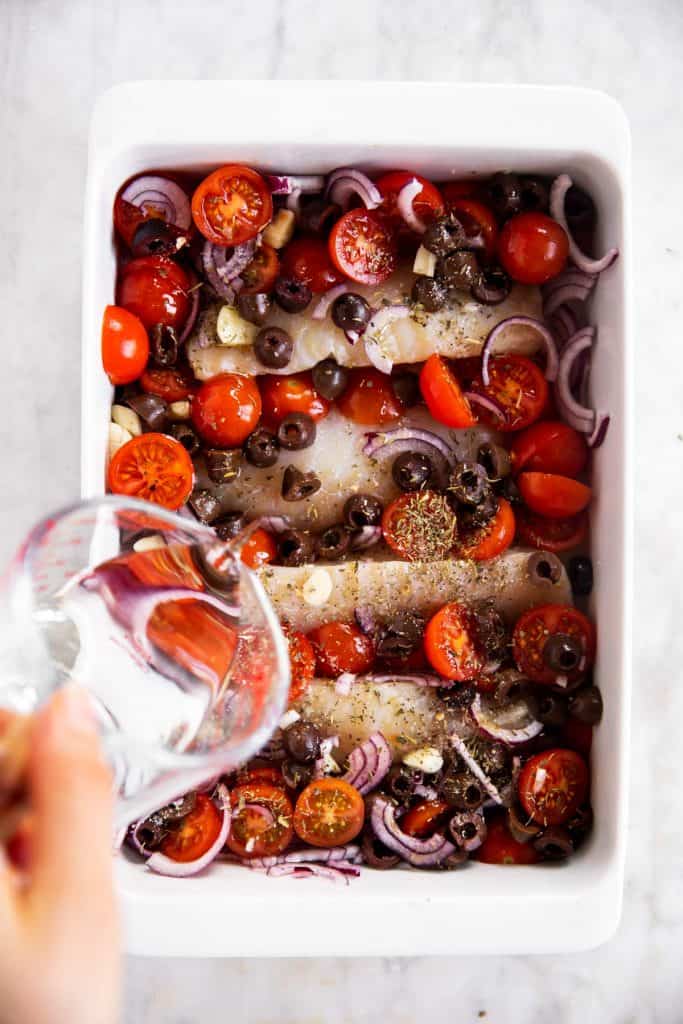 female hand pouring white wine from glass measuring jug into white dish with cod and mediterranean vegetables