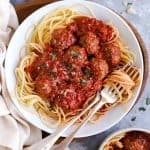 plates and bowls with italian meatballs and spaghetti, next to linen and a block of parmesan cheese