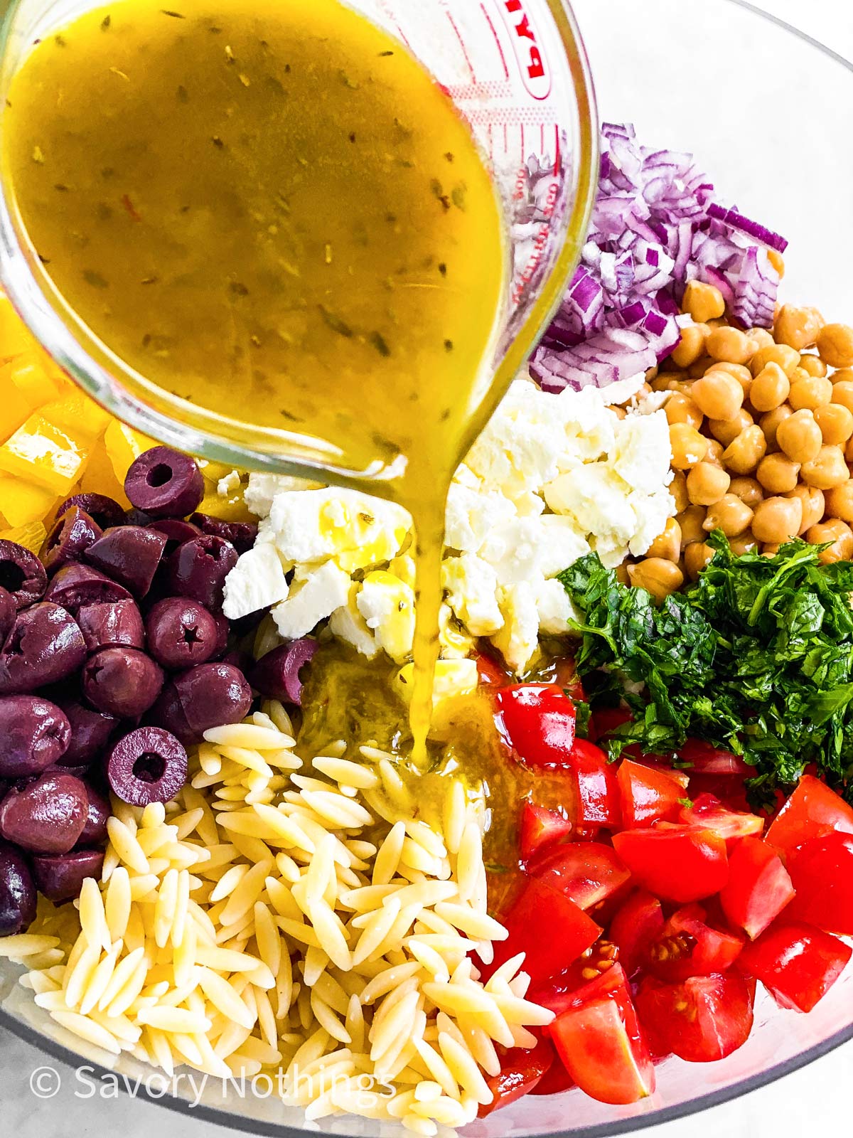 Greek salad dressing pouring over salad ingredients from glass measuring jug