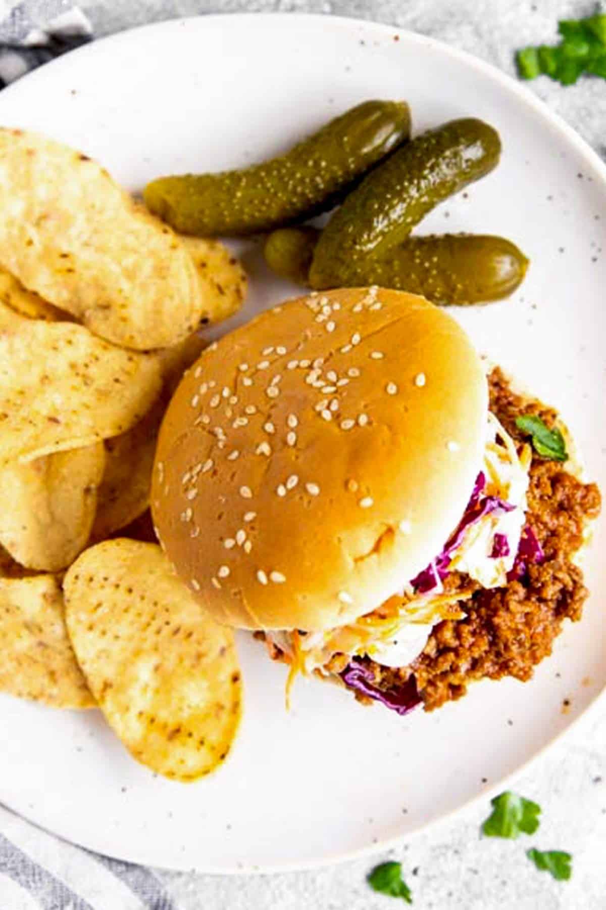 overhead view of sloppy Joe bun with chips and pickles on white plate