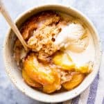 white bowl with peach cobbler, ice cream and a spoon, next to a striped tea towel