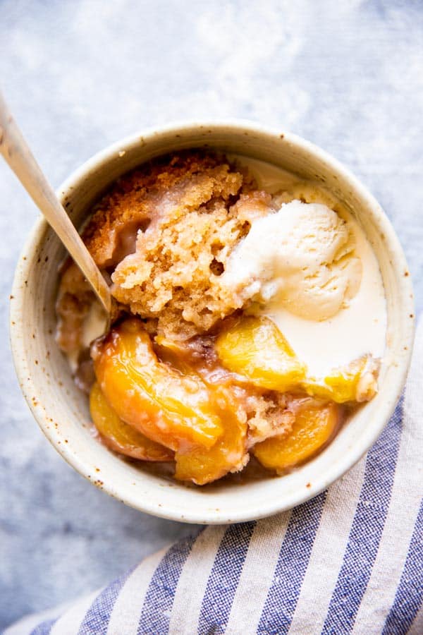 white bowl with peach cobbler, ice cream and a spoon, next to a striped tea towel