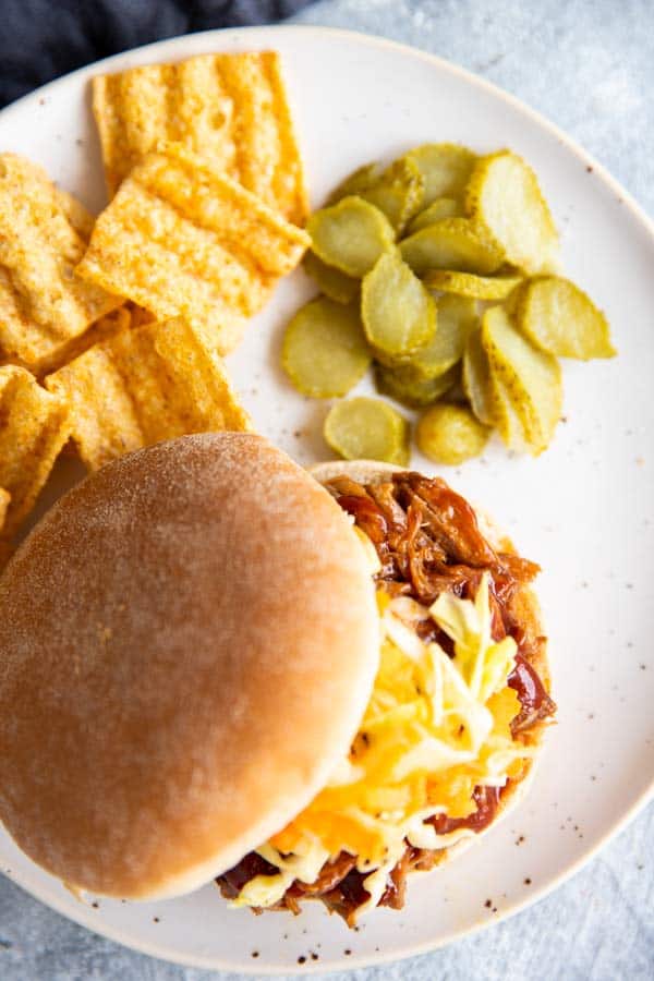 honey bbq pulled pork on a bun, on a white plate with pickles and chips