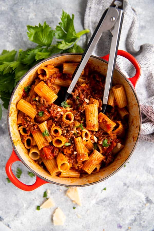 red cast iron pot with rigatoni bolognese and kitchen tongues