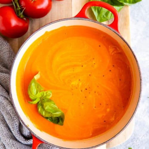 red cast iron pot with homemade tomato soup, surrounded by fresh tomatoes and basil