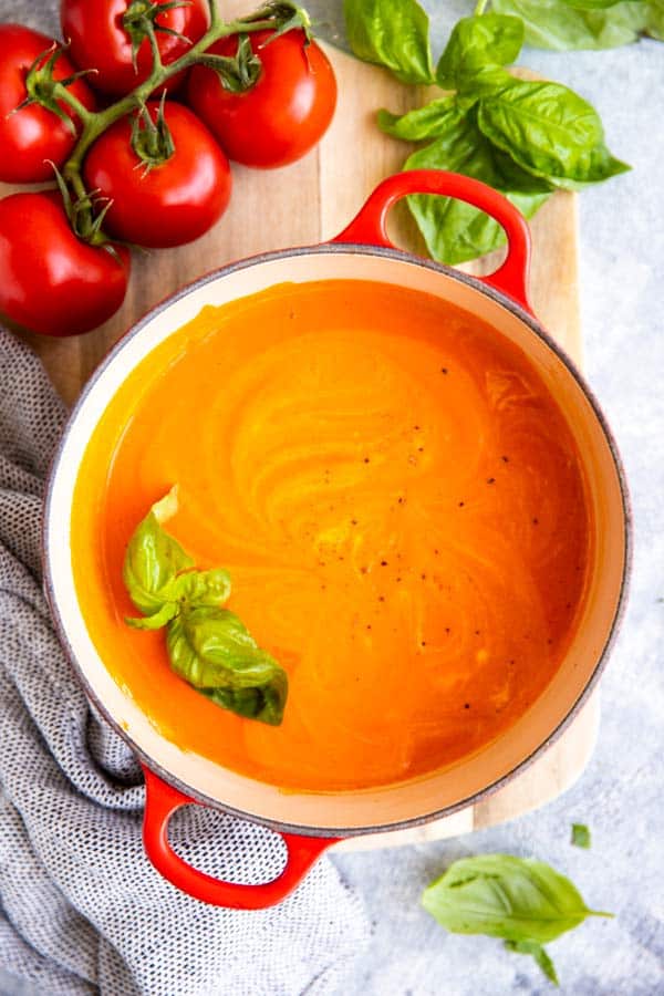 red cast iron pot with homemade tomato soup, surrounded by fresh tomatoes and basil