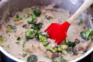 broccoli, mushrooms and chicken in creamy sauce in a sauté pan, red spatula stirring