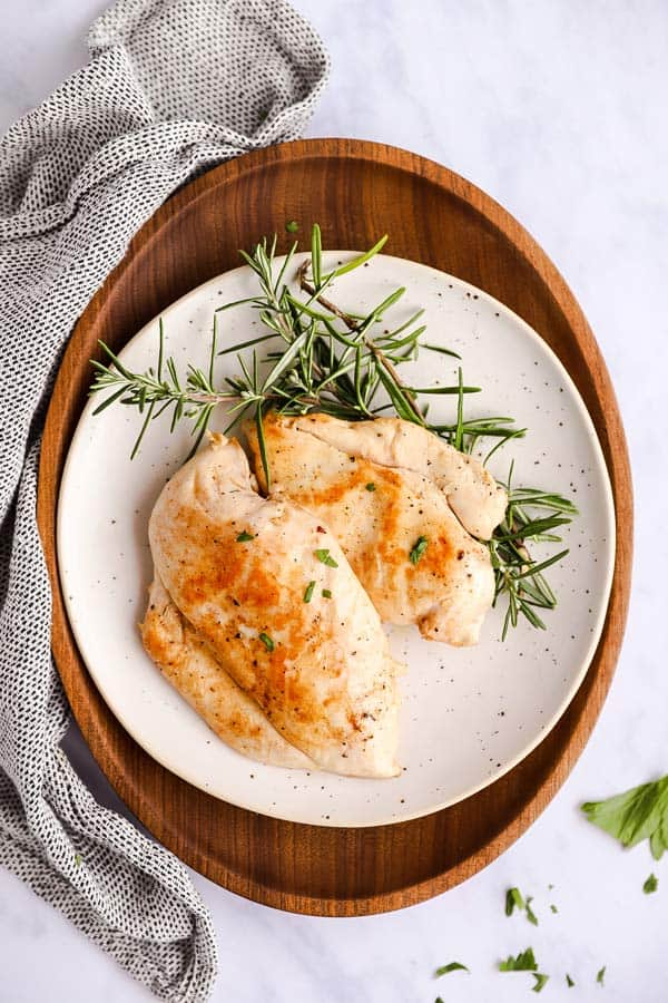 white plate on a wooden platter, with two chicken breasts and rosemary twigs on top