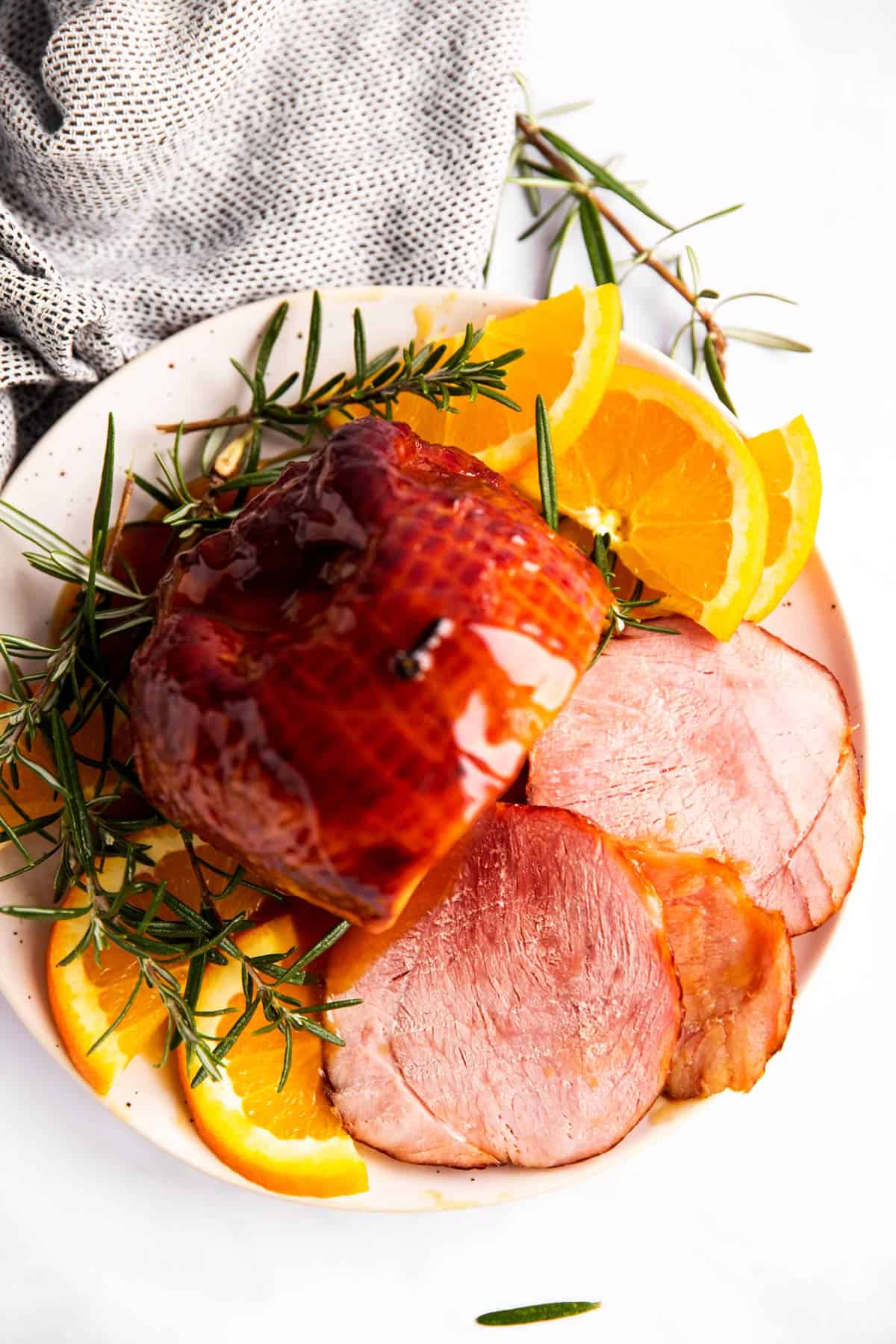 top down view of cooked ham with honey glaze on a plate with orange slices and fresh rosemary