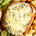vertical image of wooden board with bread bowl filled with spinach artichoke dip, surrounded by vegetables and bread